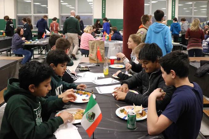 Students gathered around a table eating food and socializing in a large indoor cafeteria setting.