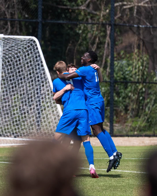Three soccer players in blue jerseys celebrate a goal. They are hugging and smiling on the field, with a soccer goal and greenery in the background.