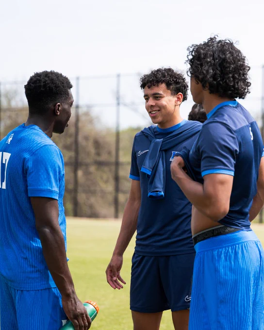 Three young men in athletic clothing converse on a sports field, with one holding a water bottle. They appear relaxed and engaged in discussion, surrounded by a black fence and greenery in the background.