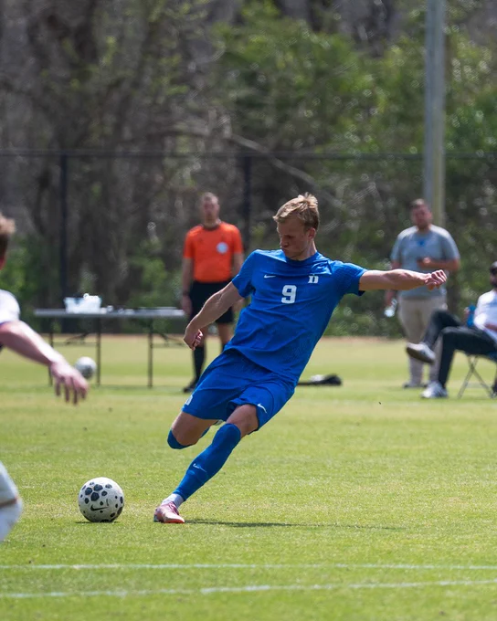 A soccer player in a blue uniform takes a shot at the ball on a grassy field. An official in an orange shirt is visible in the background, along with spectators sitting on folding chairs.