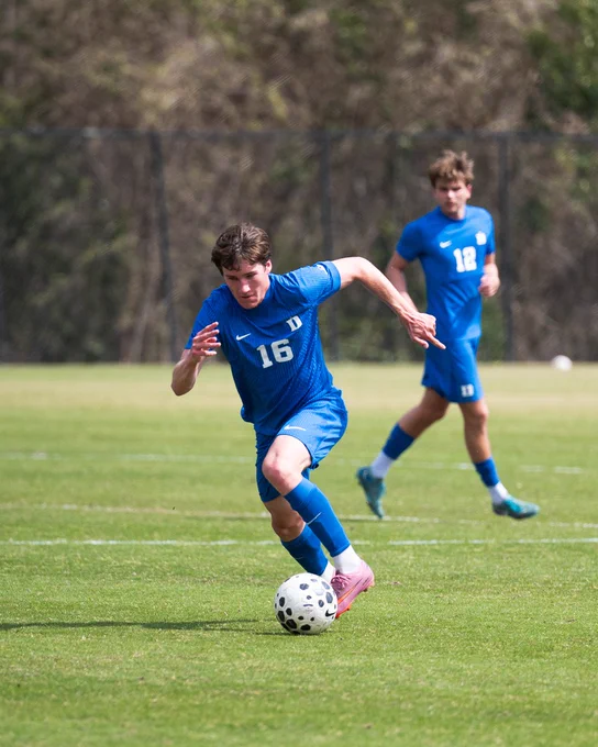 A soccer player in a blue uniform with the number 16 runs forward with the ball on a grassy field. Another player in a blue uniform with the number 12 is positioned nearby, watching the play. The background shows blurred trees and a fence.