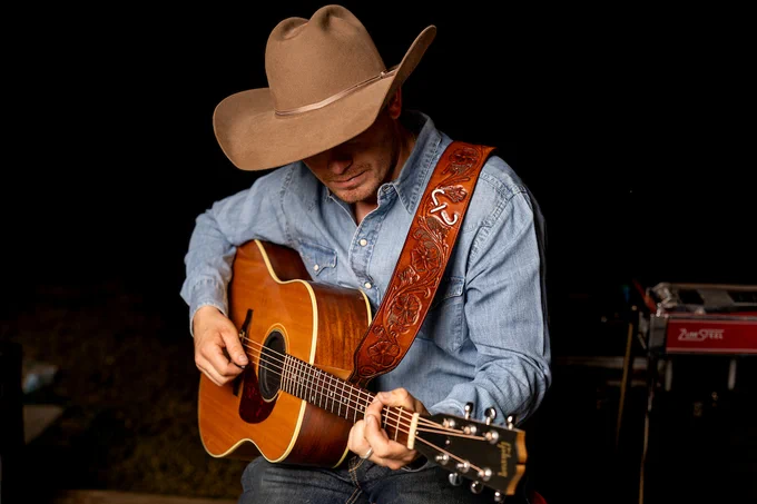 A person wearing a cowboy hat and a denim shirt is playing an acoustic guitar. The guitar has a strap with intricate designs. The background is dark, creating a focused ambiance on the guitarist.