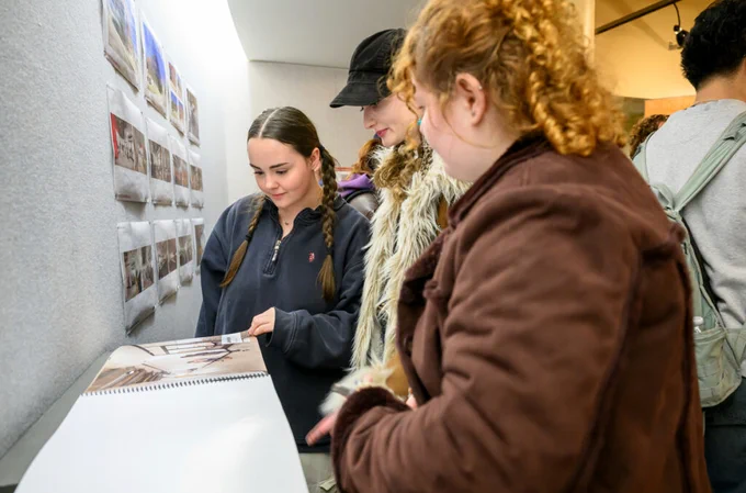 Three people are viewing photographs displayed on a wall at an exhibition. They are focused on a large open book placed on a table. The setting appears to be a gallery or similar art space.