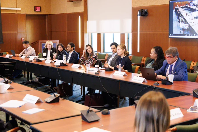 A diverse group of professionals engaged in a formal meeting around a large conference table with laptops and nameplates.