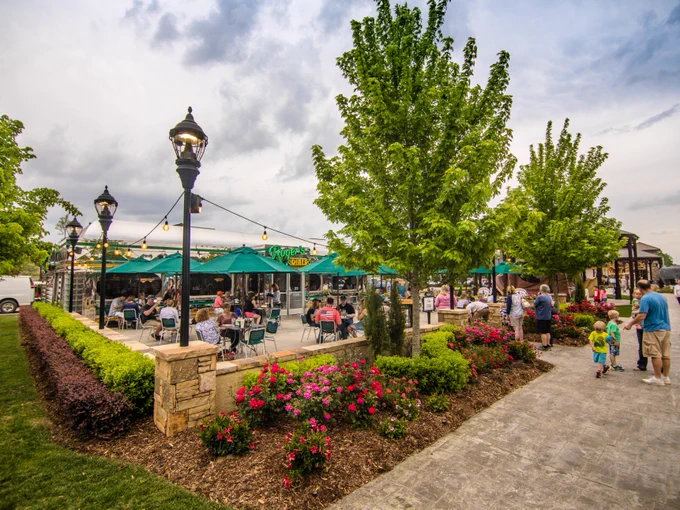 People dine outside under green umbrellas at a lively, landscaped patio surrounded by blooming flowers and trees on a cloudy day. Families and children walk along a paved path nearby.