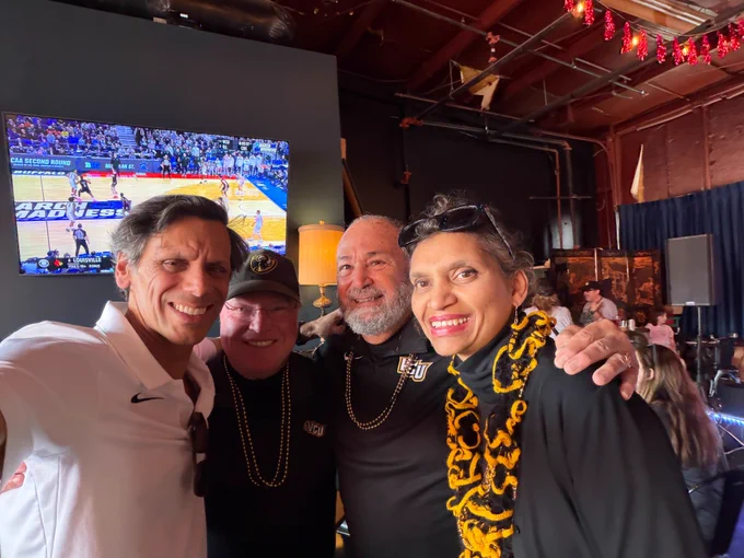 Selfie of Dr. Rao and Monica Rao smiling with two gentlemen. A basketball game plays on a large screen in the background.