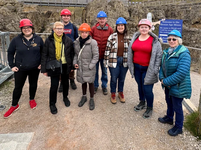 A group of eight people wearing colorful safety helmets (red, blue, orange, and pink) stand together outdoors at a rocky mining site. They are dressed in jackets and casual clothes. Behind them are rugged rock formations, a wooden walkway with railings, and a sign about a mine. The sky is overcast, and fencing lines the paths around the site.