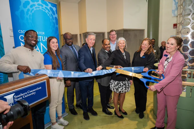 A group of people is gathered for a ribbon-cutting ceremony at Southern Connecticut State University. Blue ribbon and large scissors are used. Smiling participants and a sign with the university's name are visible in the background.