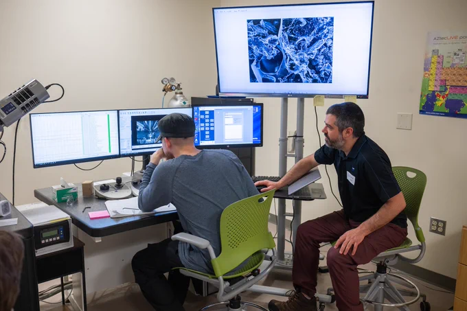 Two people are working at computer stations in a lab setting. One person is seated and looking at multiple monitors displaying data and graphs, while the other is interacting with a monitor showing a complex, magnified image on a large screen. A U.S. map decorates the wall. The environment appears focused and technical.