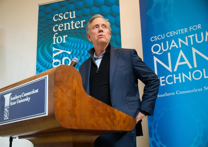 The image shows a person standing at a podium with two banners in the background. One banner reads "CSCU Center for Nanotechnology," and the other reads "Southern Connecticut State University.