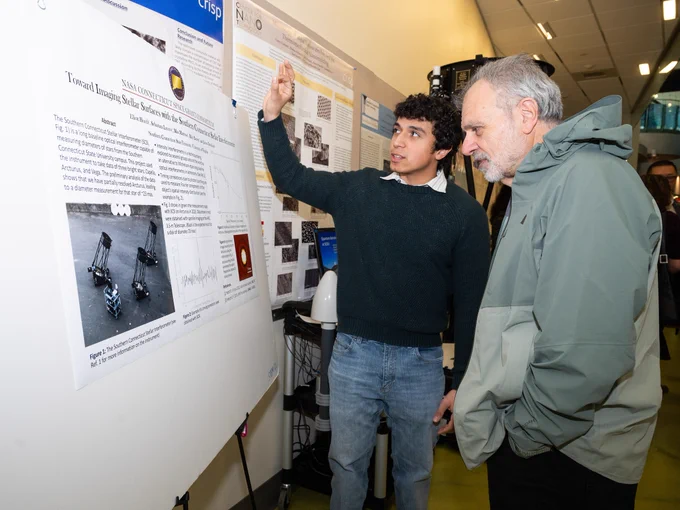 A person is presenting a research poster to another individual at an academic conference. The poster discusses solar sailing with views of sample diagrams and charts. Both individuals appear engaged in discussion, with the presenter gesturing towards the poster.