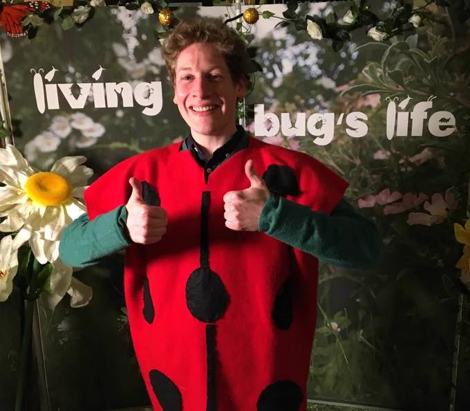 An elated Will Pearce holding his thumbs up to the camera. He is wearing a red ladybird costume while standing in front of a leafy backdrop with the text: "Living bug's life"