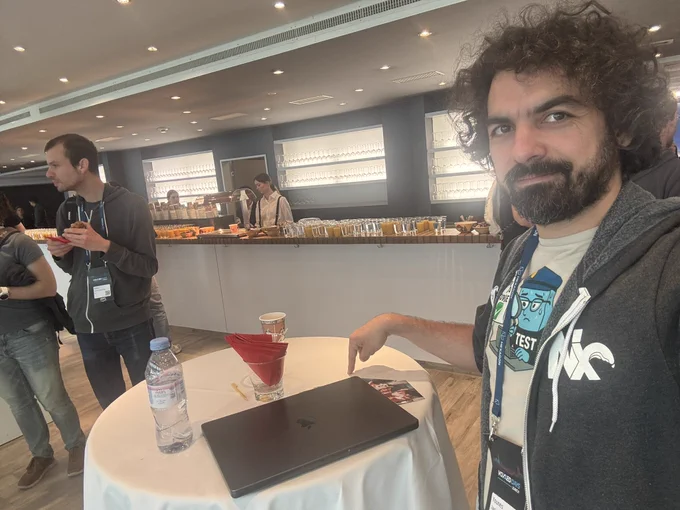 Man with curly hair and beard at a conference coffee break, pointing at a closed laptop on a small round table. In the background, attendees stand near a buffet counter with drinks and glasses.