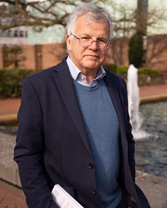 A man with short grey hair and glasses stands outdoors near a water fountain, holding documents and wearing a blazer and sweater, with trees and a brick building in the background.