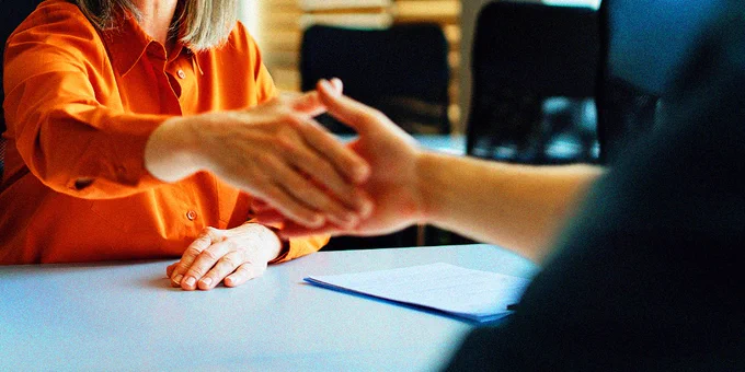 Two people shaking hands across a table in a professional office setting with documents on the table.
