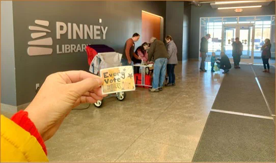Hand holding a sticker that reads 'Every Vote Matters' inside the Pinney Library lobby with voters gathered near an in-person absentee voting station in the background.