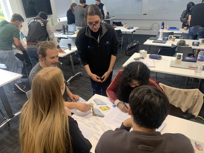 A group of students gather around a table during a hands‑on group activity as part of the Petrophysics 101 session. They are working with printed diagrams and worksheets in a classroom with a tutor providing some assistance to develop ideas and discussion. 