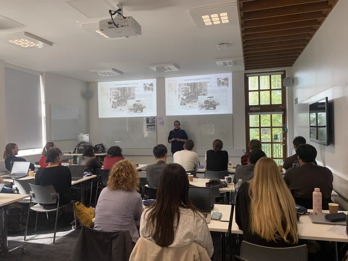 A classroom with students seated at desks facing a presenter giving a talk on the history of downhole logging. Two projected slides with historical photos and text appear at the front of the room.