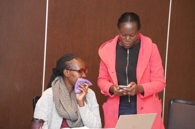 Two women are indoors in front of a wooden wall. One woman is seated, wearing glasses, a gray sweater, and a scarf, holding a purple cloth near her mouth while looking toward the other woman. The second woman stands beside her wearing a bright pink coat over a black top, focused on a smartphone she is holding. A laptop sits open on the table in front of them, along with a takeaway coffee cup, suggesting they are working or discussing something together.
