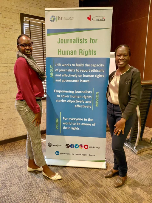 Two young ladies standing on each opposite side of a roll up banner. The banner is of the Journalists for Human Rights organization. The ladies are smiling. 