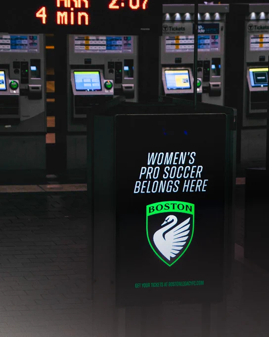A digital sign at a transit station displays the message "Women's Pro Soccer Belongs Here" above the Boston Legacy FC logo. In the background are several ticket machines.