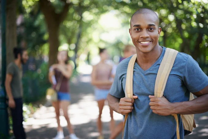 A male student smiling and wearing a backpack stands in focus on a sunlit path lined with trees. In the background, a few people are gathered, slightly blurred, suggesting a casual outdoor setting.