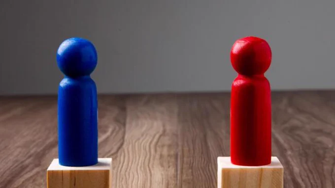 A blue chess pawn and a red pawn, each mounted on a wooden block, face one another.