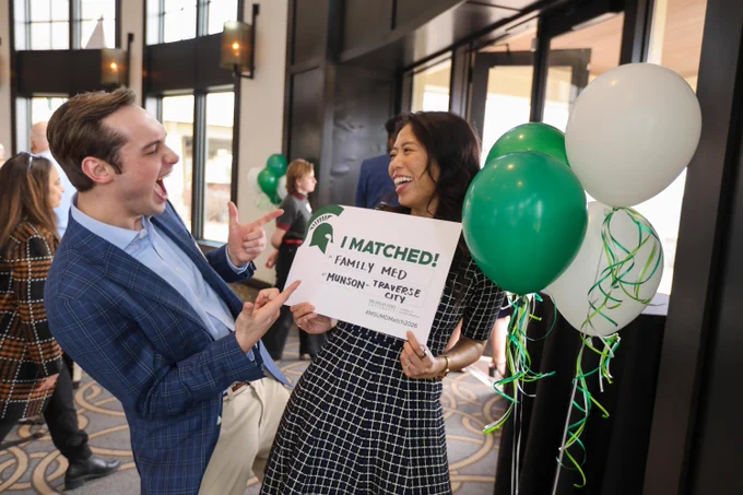 Two people laugh together while holding an \u201CI Matched!\u201D sign for a Family Medicine residency in Traverse City, with green and white balloons beside them.