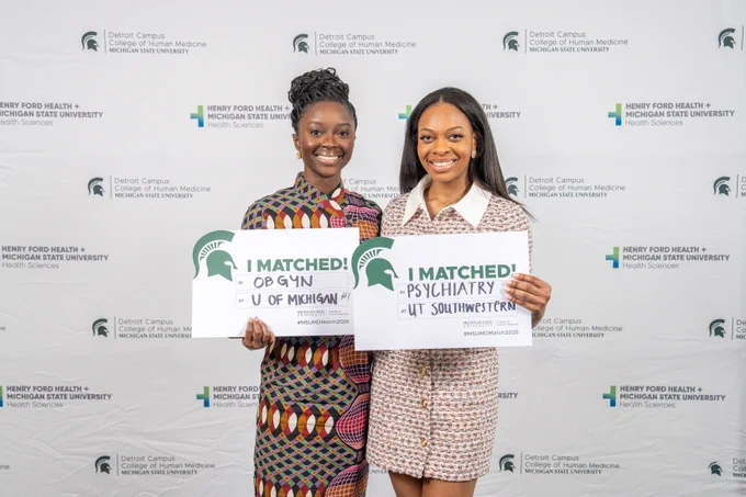 Two medical students stand in front of a Michigan State University College of Human Medicine / Henry Ford Health backdrop, smiling and holding \u201CI Matched!\u201D signs as they celebrate Match Day.