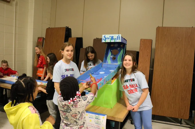 Children gathered around a homemade Sea-Ball game setup on a table in a school cafeteria.