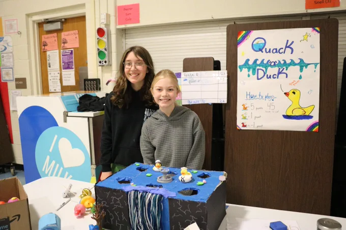 Two children stand behind a homemade 'Quack a Duck' game booth with a blue water-themed box and rubber ducks for a school event.