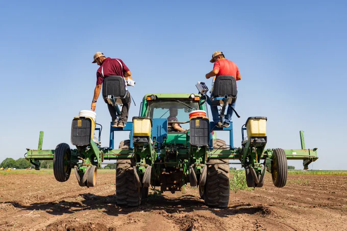 A large green John Deere tractor is in a field, with two individuals seated on its equipment in the back, performing tasks while the tractor is stationary. The sky is clear and blue.