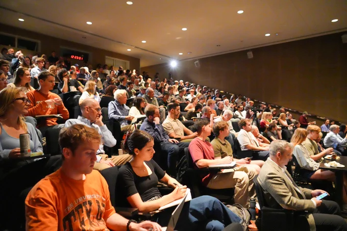 The image shows a lecture hall packed to the brim with a diverse array of individuals making up the attendee crowd for the event. They include older members of of the public and college students. 