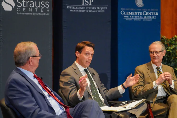 This image shows three men sitting in front of the three banners of the Strauss Center, ISP, and the Clements Center. The man in the middle gestures with his hands outwardly as he looks and speaks to the crowd; the two other gentlemen watch as he speaks. 