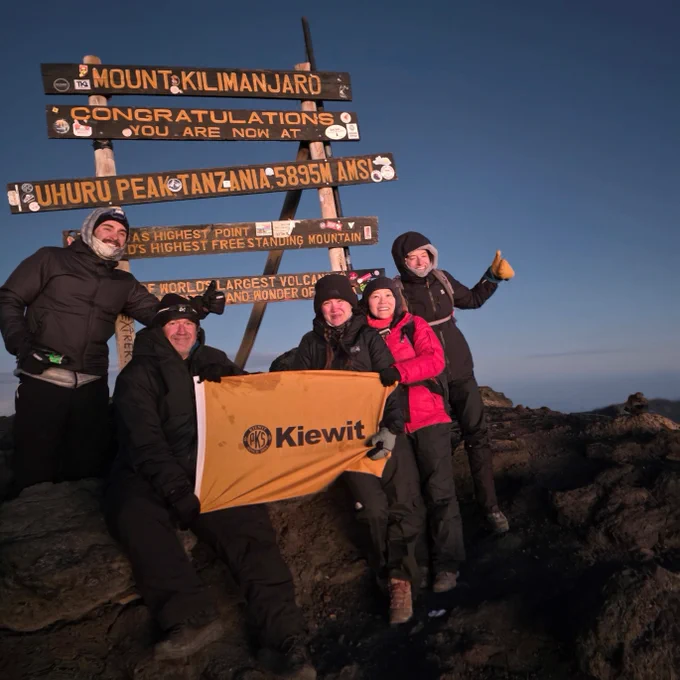 A group of people stand at the summit of Mount Kilimanjaro next to a sign reading "Congratulations, you are now at Uhuru Peak, Tanzania, 5895 M AMSL." They hold a Kiewit flag and are dressed warmly, suggesting cold weather. The sky is clear, indicating a successful climb to the top.