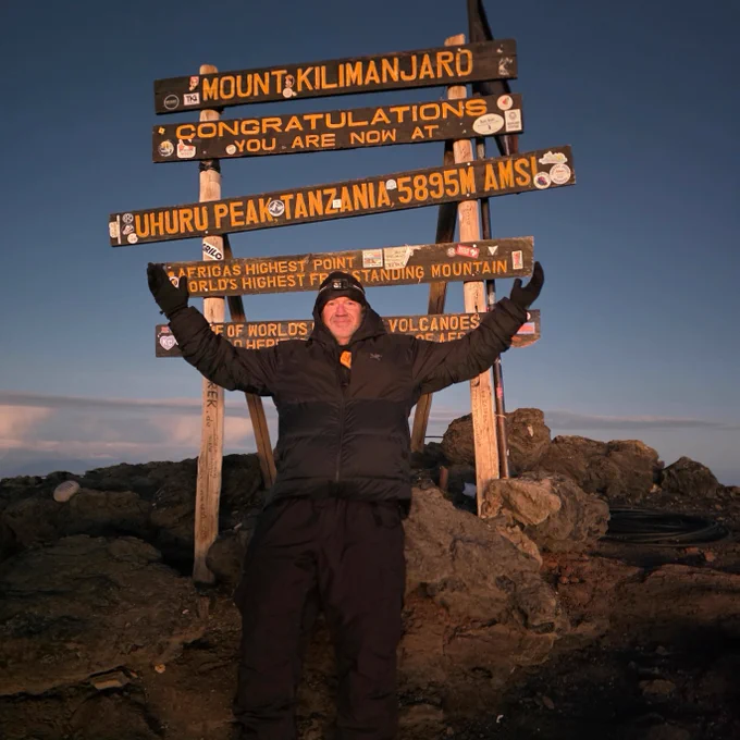 Individual standing at the summit of Mount Kilimanjaro next to a sign. The view includes rocky terrain and a twilight sky.