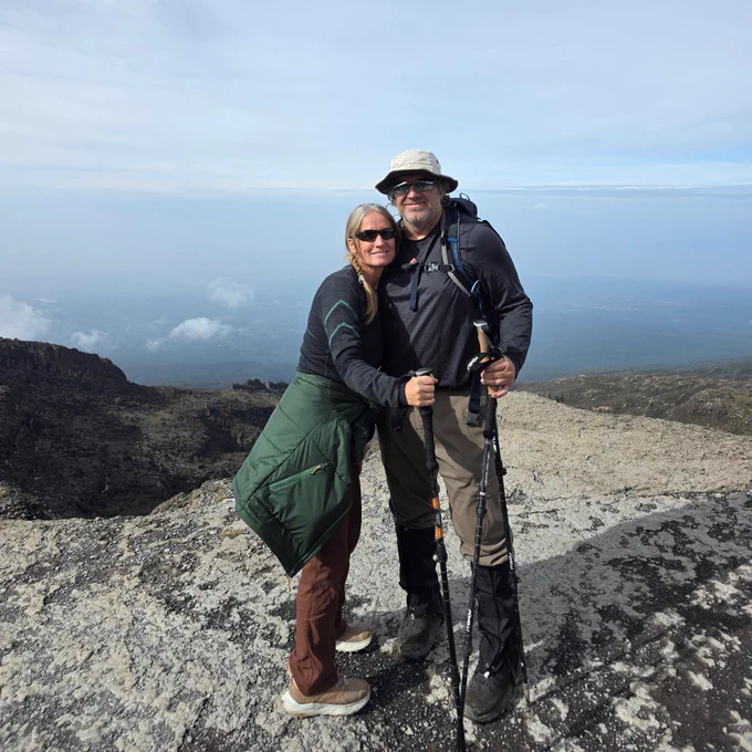 A person with long hair wearing sunglasses and a green jacket stands beside another who is equipped with hiking gear and holding trekking poles. They are both standing on a rocky terrain with a vast landscape and cloudy sky in the background.
