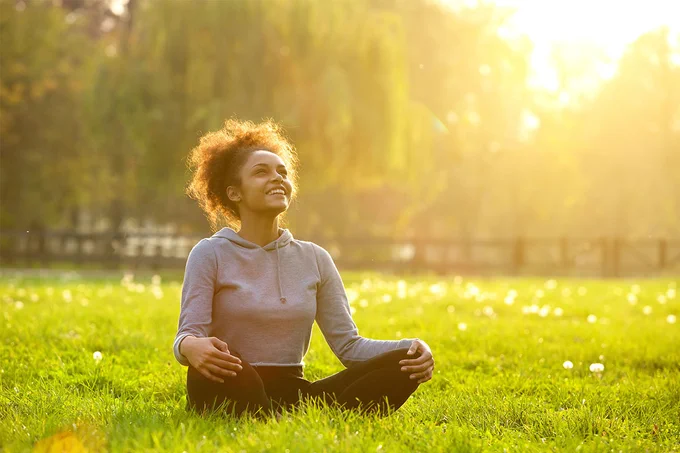 A person sits cross-legged on green grass, looking up and smiling with eyes closed. They wear a light hoodie and dark pants. The background features a blurred view of trees and a wooden fence, with sunlight creating a warm glow.