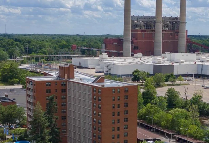 A view of REO Town in Lansing, Michigan, showing a mid-rise red brick building in the foreground, industrial buildings with three tall smokestacks in the background, and surrounding green trees under a partly cloudy sky.