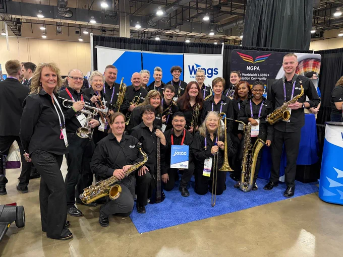 A group of people, holding musical instruments, are posing together at a conference or event. They are standing and kneeling on a blue mat, with banners for "Wings Credit Union" and "NGPA" visible in the background.