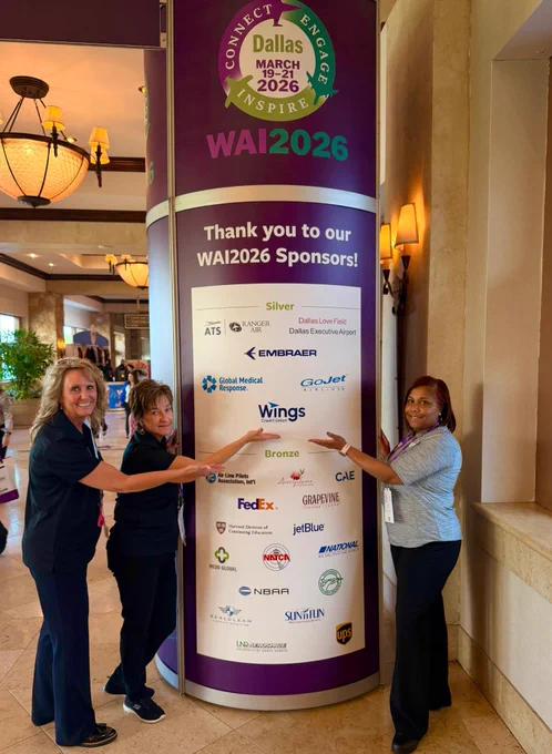 A group of three people is posing around a tall display banner for the WAI2026 event in a hotel lobby. The banner lists several sponsors, including FedEx, Boeing, and CAE. The background includes a chandelier and other decorative elements.