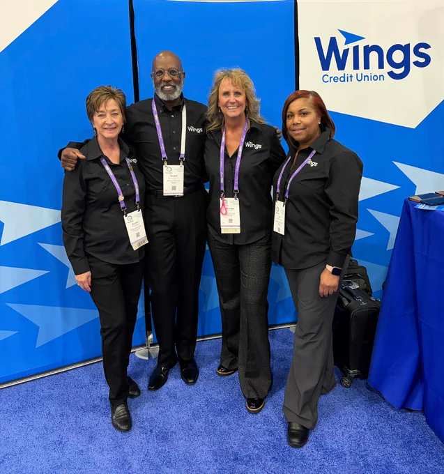 A group of four people stands in front of a Wings Credit Union backdrop, wearing matching outfits with name tags and conference lanyards. They are smiling and appear to be at a professional event.