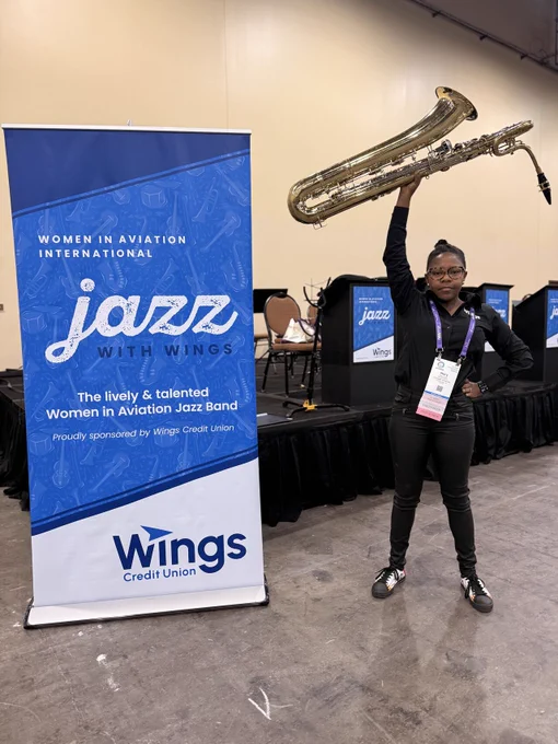 Individual standing beside a banner for "Women in Aviation International Jazz with Wings," lifting a large saxophone overhead. The event is sponsored by Wings Credit Union. Stage setup with chairs and a jazz-themed backdrop visible in the background.