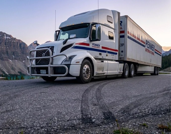 A white Penner International transport truck with blue and red stripes is parked on a mountain highway pull off at sunrise. The truck is positioned slightly from the side, showing the cab, the trailer, and tire marks on the pavement. Tall rocky mountains and a faintly glowing sky create a scenic backdrop.