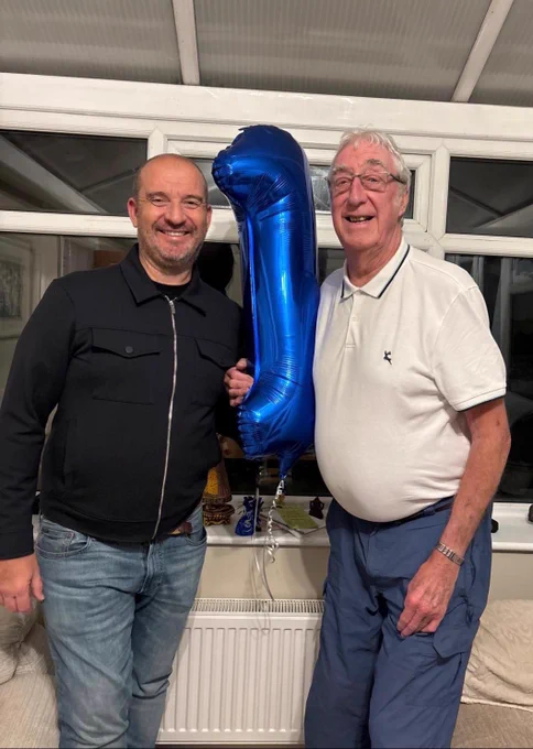 Ian Tonks (left) and Graham Sisson (right) standing indoors holding a large blue balloon shaped like the number one.