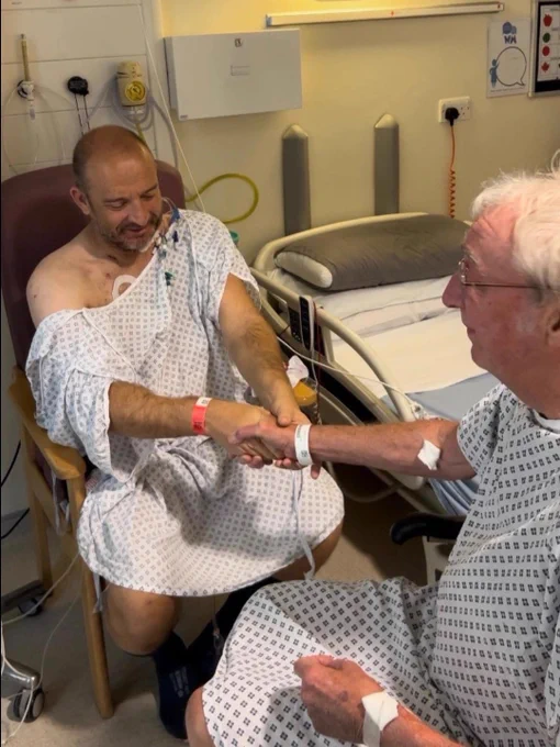 Ian Tonks (left) and Graham Sisson (right) in hospital gowns holding hands before surgery, seated beside a hospital bed 