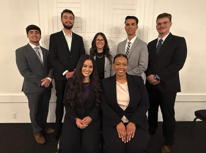 A group of seven people dressed in professional attire, standing and seated indoors against a neutral wall backdrop. They are smiling and appear to be at a formal or business event.