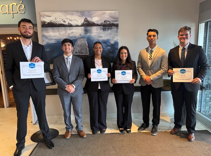 A group of six people standing indoors, dressed in business attire, holding certificates. Behind them, a large photograph of a wooden pier extending into a body of water with snow-capped mountains in the distance is displayed.