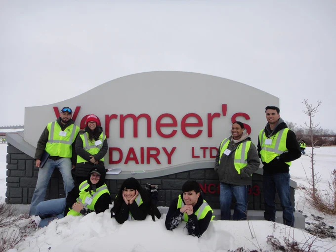 A group of individuals wearing high-visibility vests pose in front of a sign reading "Vermeer's Dairy LTD." The ground is covered in snow.