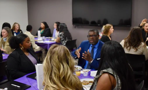 A group of people sit around tables in a meeting room, engaged in conversation during a meal. The tables are covered with purple tablecloths, and there is a large screen on the wall.